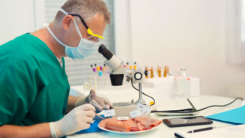 masked and gloved man looking at processed meat under microscope