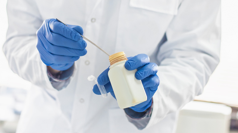 person in white lab coat with blue gloved hands dipping test strip into bottle