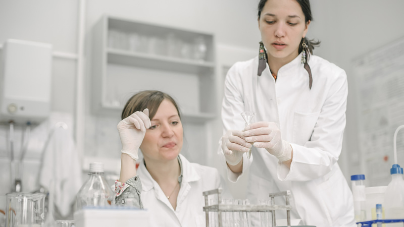2 women lab techs in white lab coats and gloves with glass beakers
