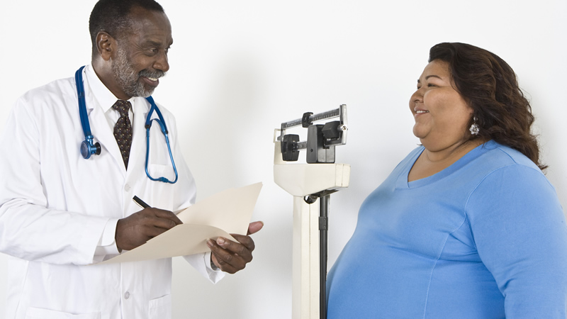 doctor writing in folder, obese woman standing on weigh scale
