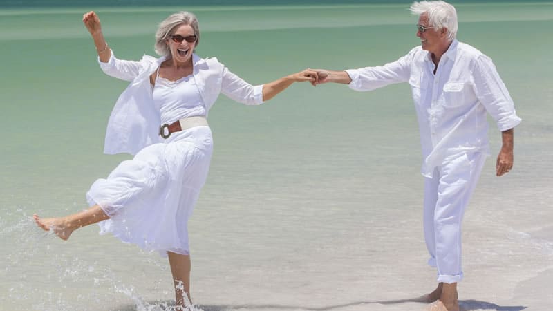 smiling older couple with white hair, in white clothes on beach dancing