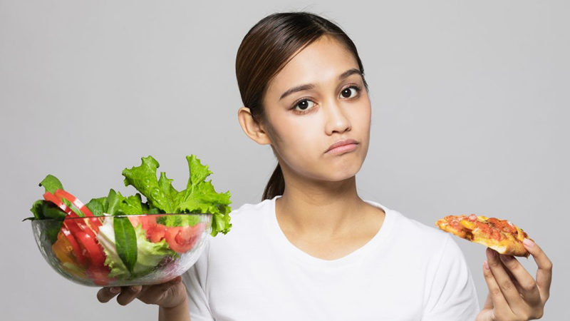 woman holding large salad and slice of pizza as if deciding which