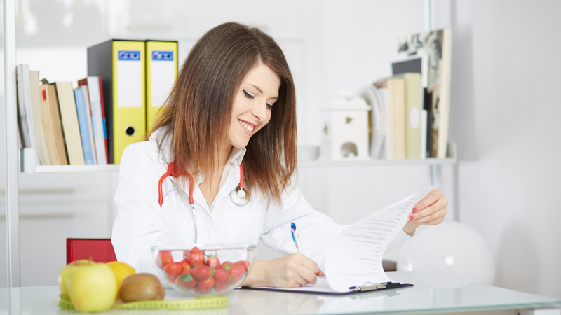 smiling woman in white lab coat and stethoscope writing on clipboard, fruit and measuring tape on desk