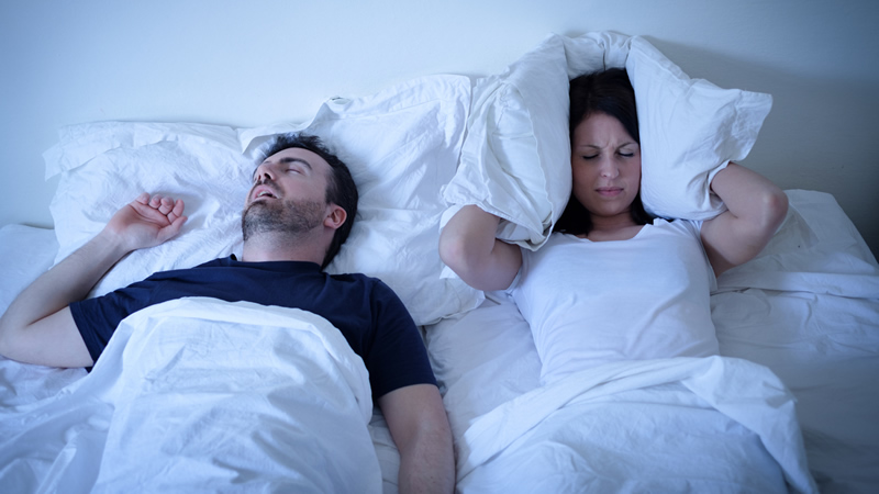 couple in bed, man with mouth open as if snoring, frowning woman holding her pillow to cover her ears