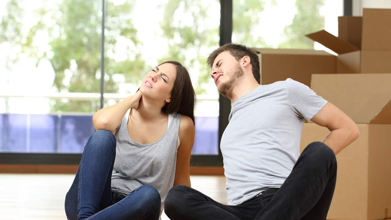 young couple sitting on floor, wincing in pain with moving boxes behind