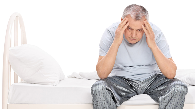 older man sitting on edge of bed holding his head as if in pain