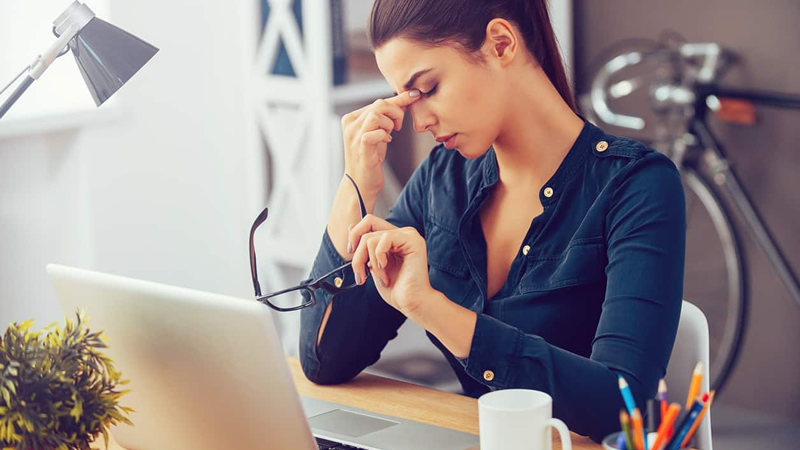 woman at desk holding her nose between her eyes as if tired in pain