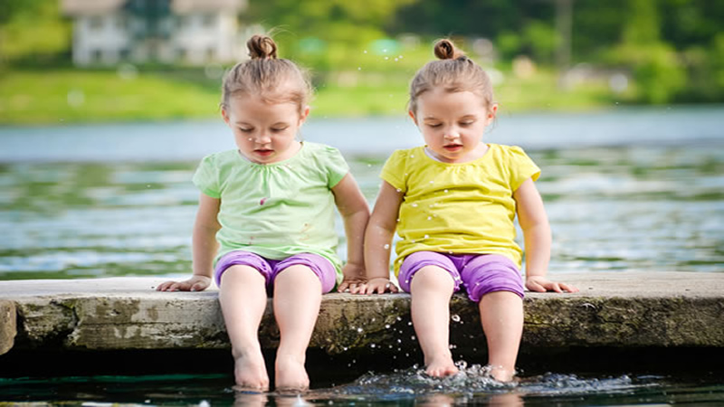 young twin girls sitting on dock dangling their feet in lake water