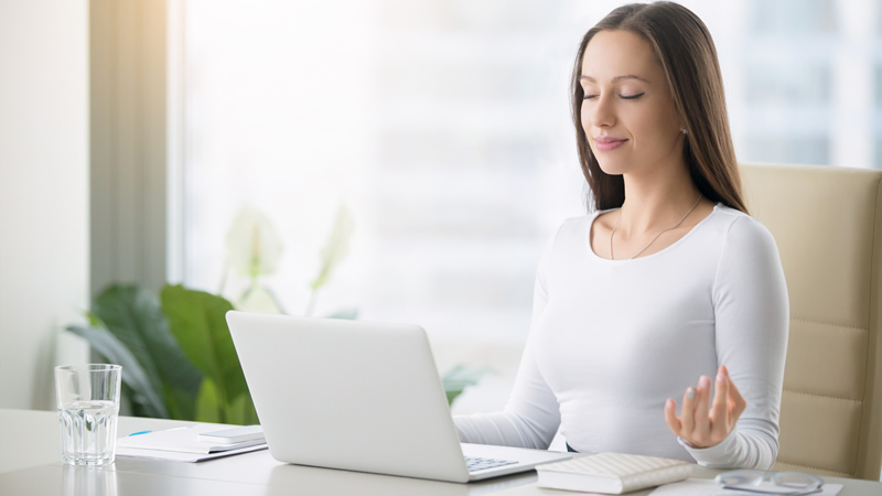 smiling woman at desk with eyes closed and hands in meditation pose