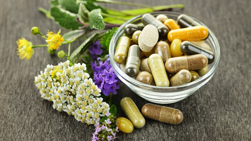 glass bowl of herbal tablets and capsules, surrounded by herbal flowers