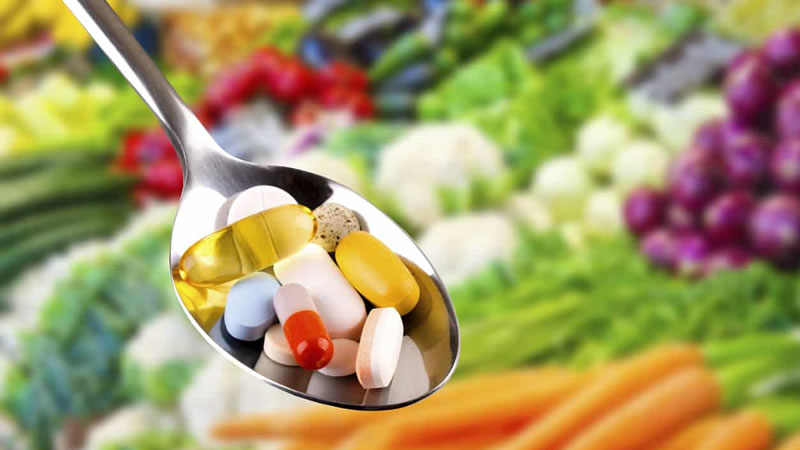 tablets and capsules on silver spoon, vegetables behind
