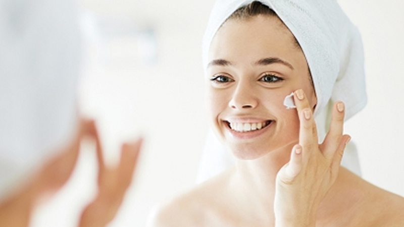 young smiling woman with hair in white towel applying cream to her face