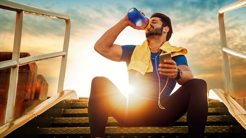 athletic man sitting on stairs, earbuds, towel on shoulders, drinking water, sunshine in back